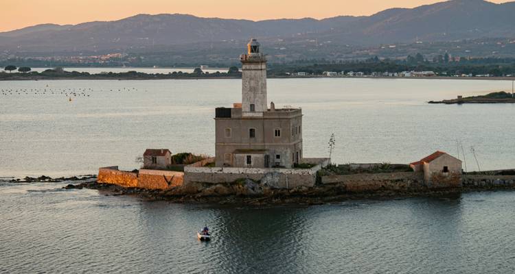 Ein verwitterter Leuchtturm auf einem kleinen felsigen Eiland leuchtet im warmen Abendlicht, während ein winziges Boot in der Nähe kreist.