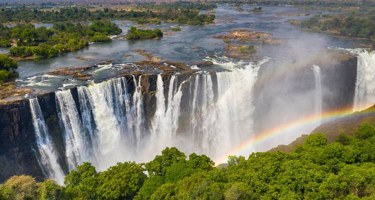 Luftaufnahme der Viktoriafälle mit Regenbogen über der nebelverhangenen Schlucht und üppigen Flussinseln stromaufwärts.