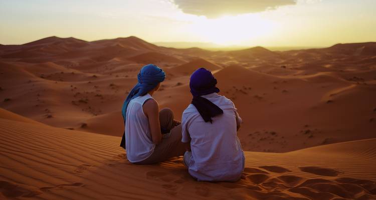 Zwei Reisende in blauen Turbanen sitzen auf einer hohen Düne und beobachten den Sonnenuntergang über endlosen Sahara-Sandwellen.
