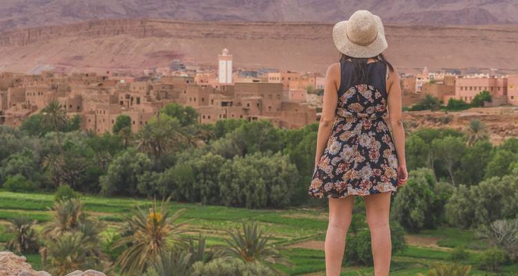 Eine Frau in einem geblümten Kleid und Sonnenhut blickt auf eine üppige Oasenstadt mit einem weißen Minarett, das von trockenen Bergen im Hintergrund umrahmt wird.