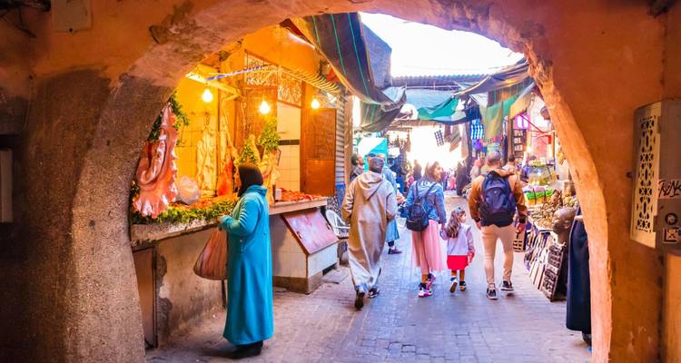 Callejón de mercado lleno de gente bajo un arco con luces coloridas, puestos y compradores en ropa tradicional