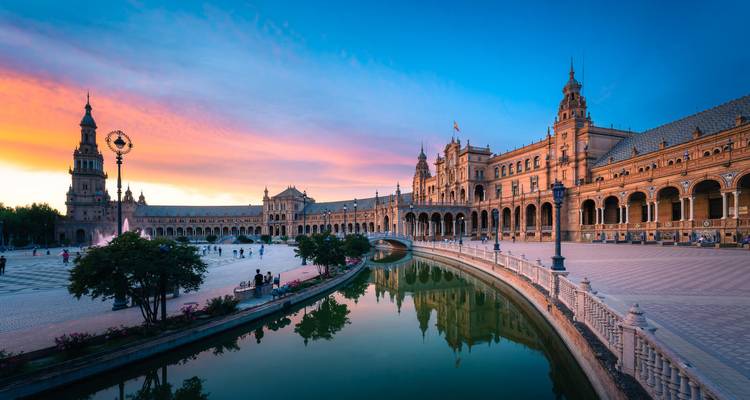 Der geschwungene Palast der Plaza de España, der Kanal und der leuchtende Sonnenuntergangshimmel spiegeln sich im ruhigen Wasser.