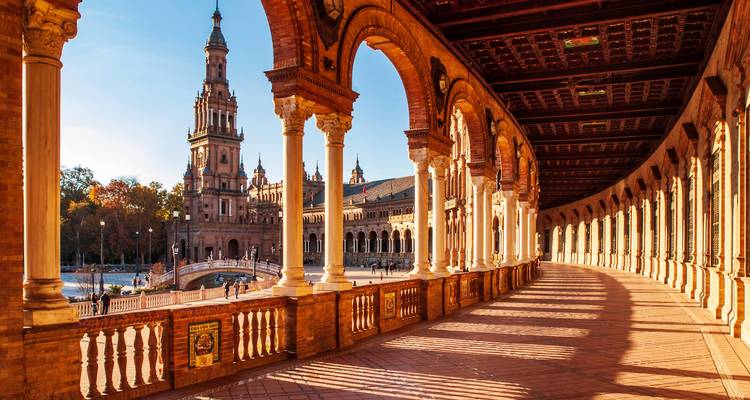 Sonnenbestrahlter gewölbter Korridor der Plaza de España mit langen Schatten und Turm in der Ferne.