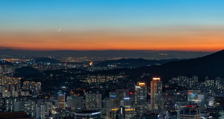 Schemering stadsgezicht van Seoel met verlichte wolkenkrabbers, bergen op de achtergrond en een dunne wassende maan boven de horizon
