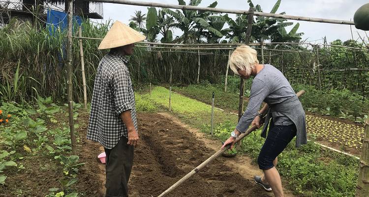 Reiziger helpt lokale boer met het bewerken van grond op een klein groenteplantje omgeven door bananenbomen.