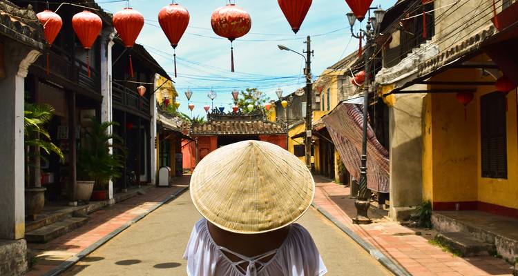 Achteraanzicht van reiziger in een punthoed die tussen rode lantaarns door slentert langs de historische straat van Hoi An.