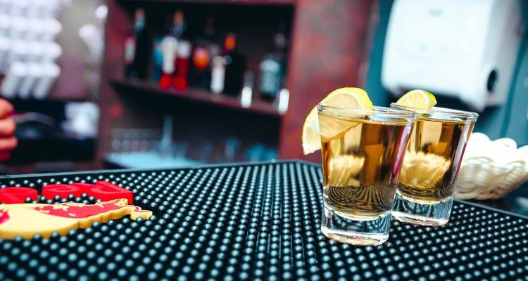Zwei Tequila-Shots mit Zitronenscheiben auf einer schwarzen Barmatte in einer schwach beleuchteten Bar