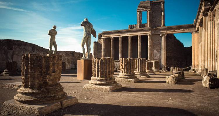 Ancient columns and bronze statues amid the sun-lit ruins of Pompeii against a clear blue sky.