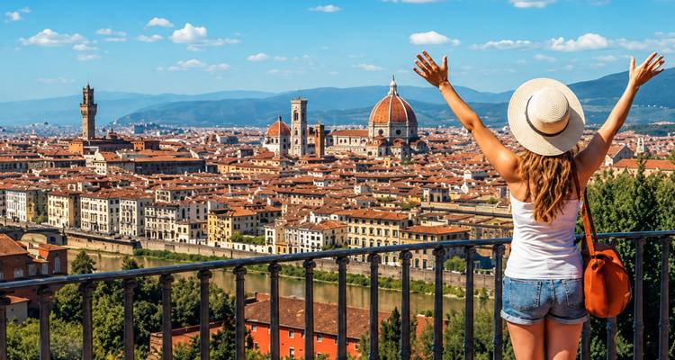 Woman in sunhat joyfully raising arms at a viewpoint overlooking Florence’s terracotta rooftops and iconic Duomo.