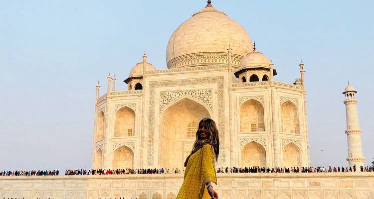 A woman in a yellow dress poses cheerfully in front of the Taj Mahal at sunrise.