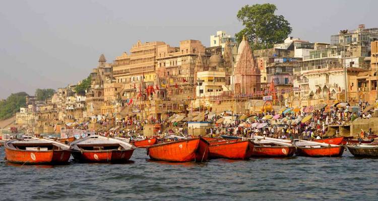 Ikonische Flussuferpromenade von Varanasi mit farbenfrohen Ghats, Tempeln und roten Ruderbooten auf dem Wasser.