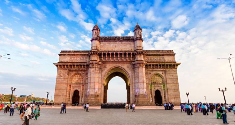 The Gateway of India monument framed by a dramatic blue sky with crowds walking on the waterfront plaza.