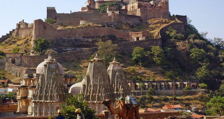 Camel riders passing ornate Jain temples below the massive ramparts of Kumbhalgarh Fort on a sunny day.