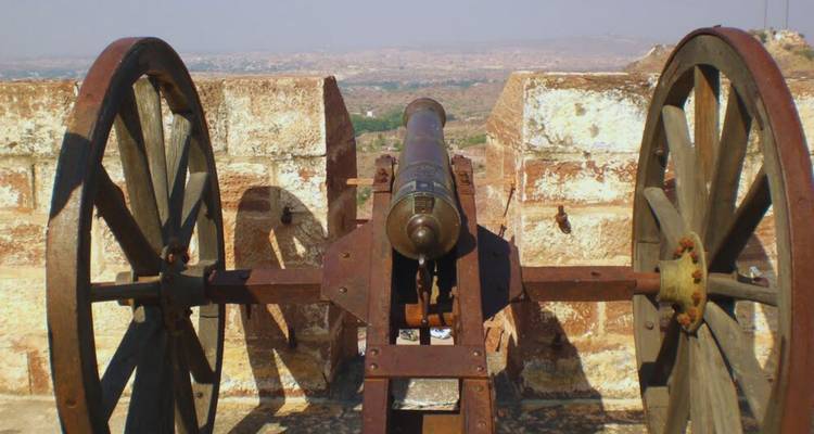 Old rusted cannon aimed over sandstone battlements with hazy desert landscape beyond.