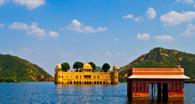 Jal Mahal water palace reflected in the blue waters of Man Sagar Lake, framed by green hills and clouds.