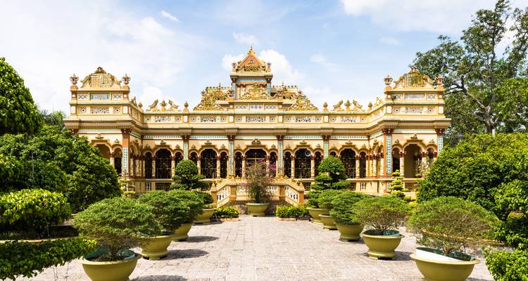 Intricately decorated Vinh Trang Pagoda with golden stucco and manicured bonsai trees under bright sky.