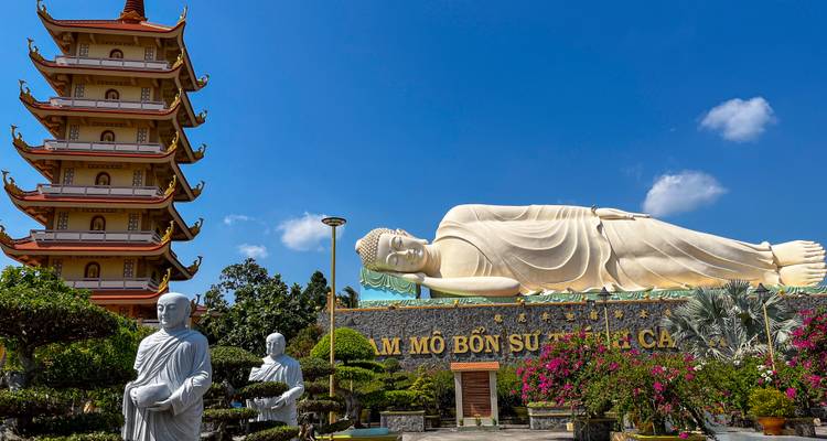 Reclining Buddha statue behind manicured bonsai and multi-tiered pagoda under bright blue Mekong Delta sky.