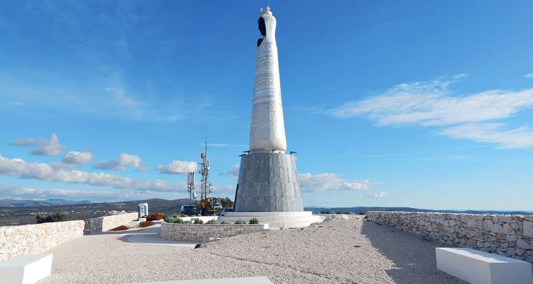 Tall modern Marian statue atop a cylindrical plinth on a rocky hilltop viewpoint against a vast blue sky.