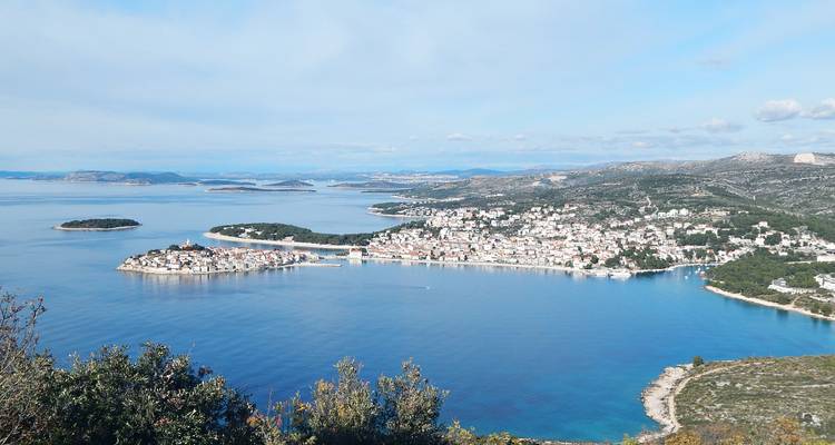 Panoramic view of a small fortified peninsula town jutting into vivid blue waters surrounded by tiny islands and distant mountains.