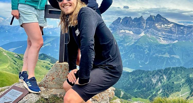 Wanderer an einer Gipfelmarkierung mit Blick auf dramatische Alpengipfel und Täler.