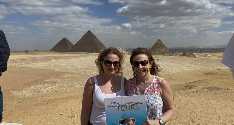 Twee vrouwen poseren met een tourbord voor de Piramides van Giza onder een deels bewolkte hemel.