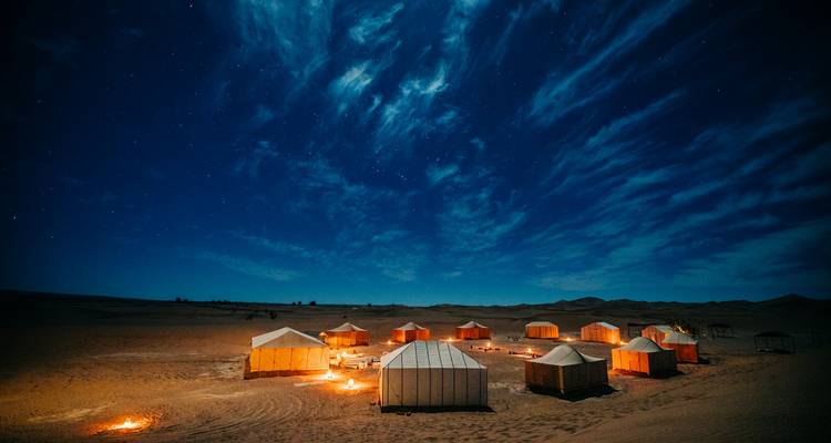 Glowing desert camp tents encircle small fires under a brilliant star-filled night sky.