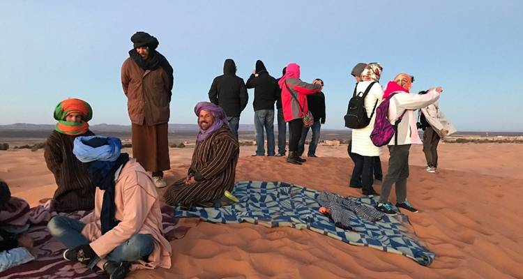 Travelers wrapped in scarves and jackets gather on a dune at dawn with blankets spread on the sand.