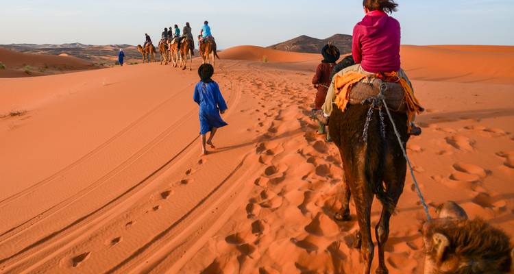 Line of tourists riding camels across orange dunes led by a blue-robed guide and a child walking barefoot.
