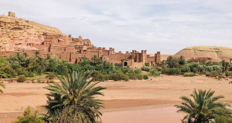 Earthen ksar of Ait Benhaddou rises above a dry riverbed framed by palms and desert hills.