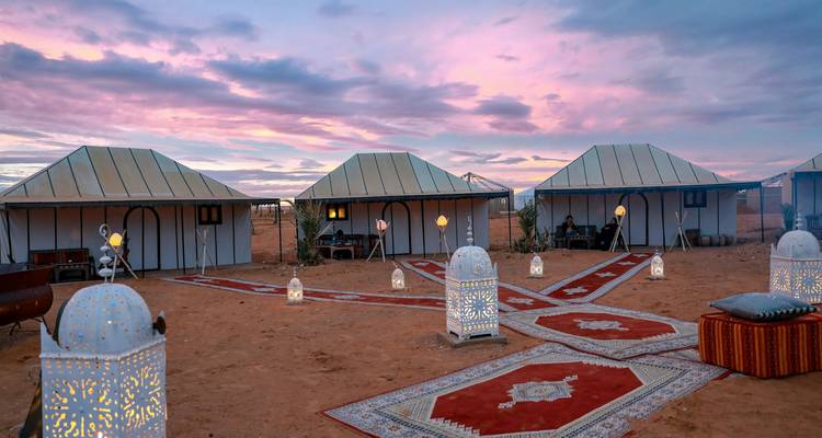 Chic desert camp with patterned carpets, lanterns and white canvas tents beneath a pink dawn sky.