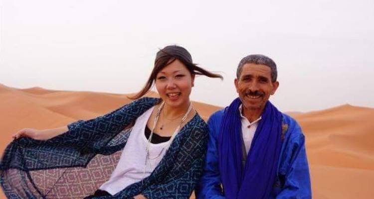 Smiling traveller with local Berber man seated on dunes.