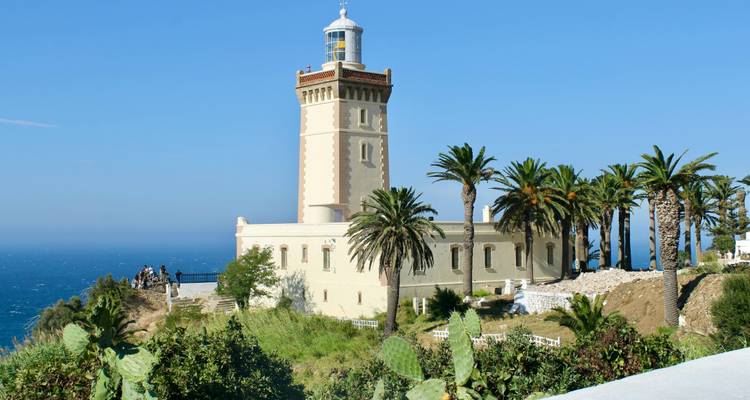 Cream-colored lighthouse perched on a cliff surrounded by palms overlooks the Atlantic Ocean.