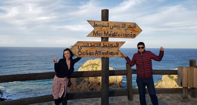 Happy couple pose by wooden sign pointing to the Mediterranean and Atlantic with coastal cliffs behind.