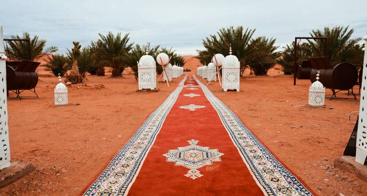 Long red carpet flanked by ornate lanterns leads to a desert camp among palms.