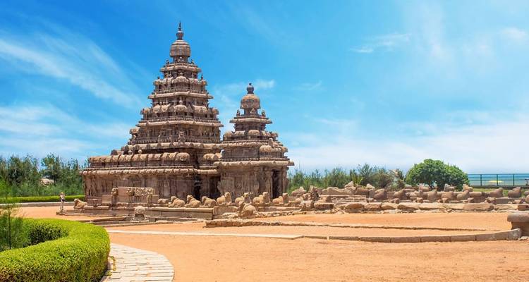 Shore Temple of Mahabalipuram stands against a bright blue sky surrounded by sandy grounds.