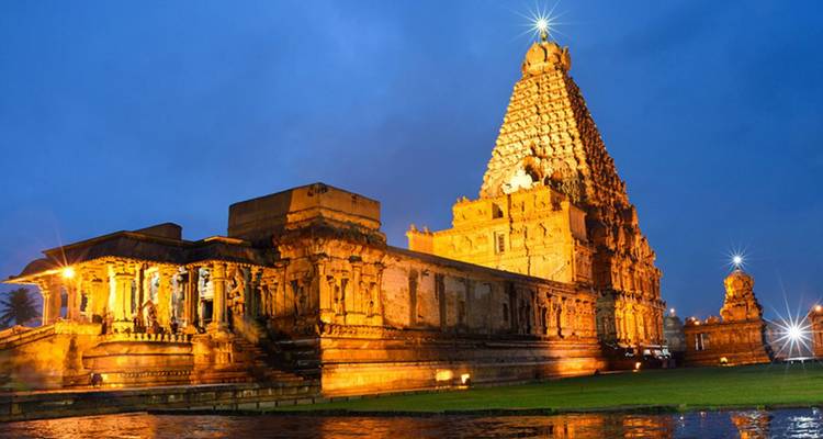 Illuminated Brihadeeswarar Temple reflects in water at twilight, showcasing towering architecture.