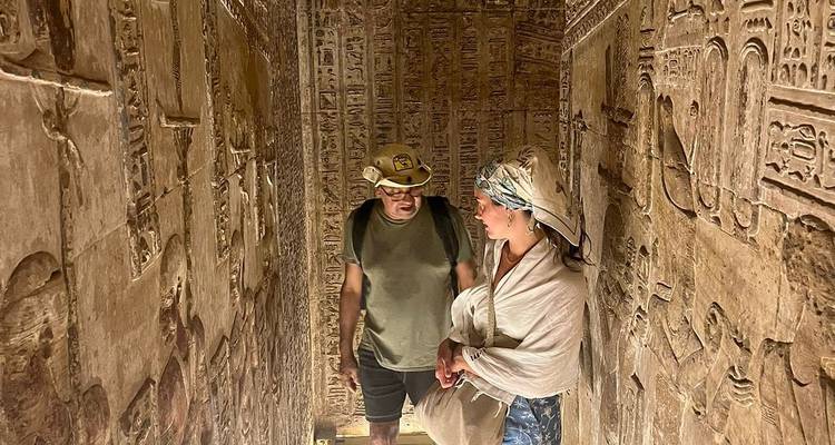 Twee reizigers bekijken gedetailleerde muursculpturen in een smalle, zwak verlichte doorgang van een oude tempel.