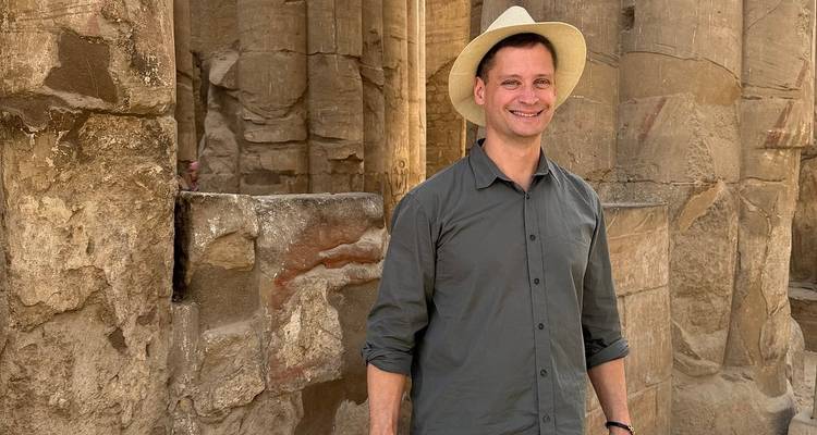 Een man in een lichte hoed glimlacht terwijl hij tussen verweerde zandstenen zuilen van een Egyptische tempel staat.