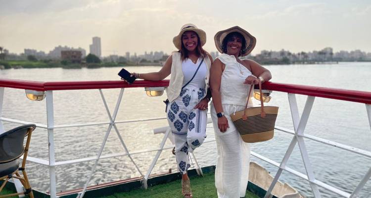 Deux femmes souriantes vêtues de tenues d'été posent sur le pont découvert d'un bateau fluvial avec le Nil et la ligne d'horizon du Caire en arrière-plan.