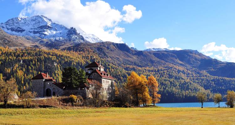 Manoir de pierre à côté d'un lac alpin turquoise avec des mélèzes d'automne colorés et des pics enneigés au-delà.