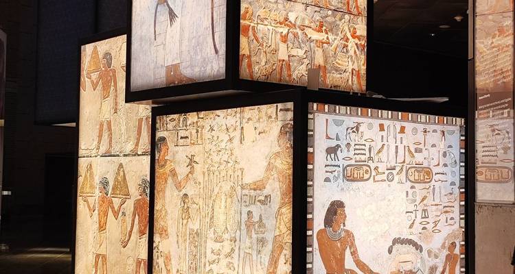 Des cubes de musée illuminés affichant des peintures murales égyptiennes anciennes colorées et des hiéroglyphes dans une galerie sombre.