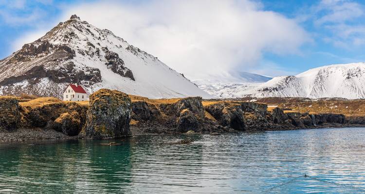 Eenzaam wit huisje met rood dak naast een turquoise baai en een met sneeuw bedekte berg op de achtergrond.