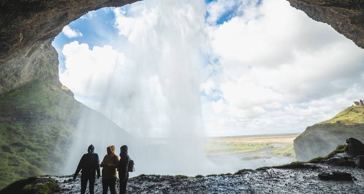 Bezoekers in silhouet kijken naar de waterval vanaf achter het bruisende water, omlijst door de grotteopening die uitkijkt op open vlaktes