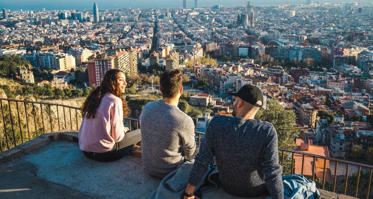 Drei Freunde sitzen an einem Aussichtspunkt auf einem Hügel und blicken auf die sich ausbreitende Stadt Barcelona hinunter