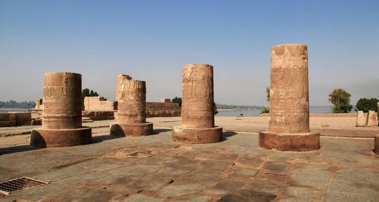 Rangée de colonnes en grès usées par le temps se dressant sur une cour en pierre à côté du Nil sous un ciel bleu dégagé.