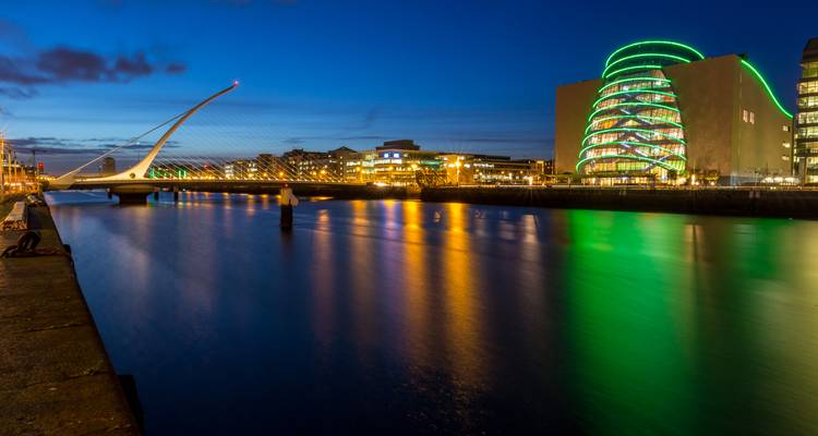 Nächtliche Flussfront-Szene in Dublin mit beleuchteter Brücke und neongrünem Kongresszentrum, das sich im ruhigen Wasser spiegelt.