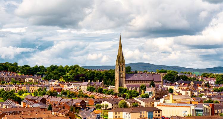 Panoramique urbain de Londonderry présentant une haute flèche d'église contre des collines vertes ondulantes et des nuages dramatiques.