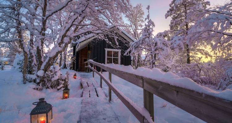 Snow-covered cabin nestled among frosted trees at twilight with lanterns lighting a wooden walkway.