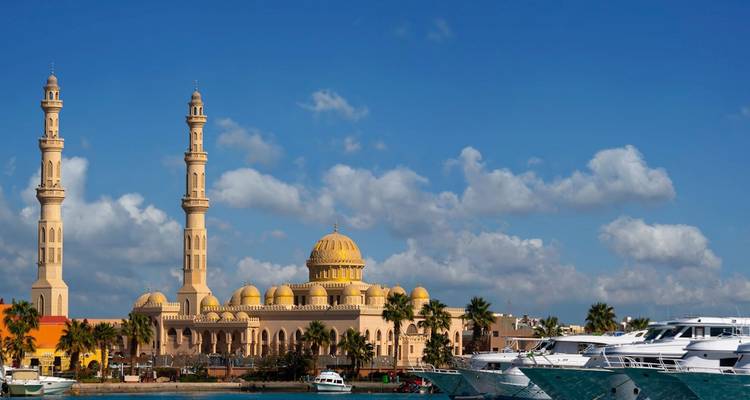 Grote moskee met gouden koepels en dubbele minaretten die uitkijken op de jachthaven vol jachten onder een blauwe hemel.