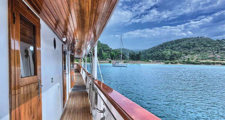 Wood-trimmed deck corridor of cruise yacht overlooking calm blue Adriatic waters and green hills.